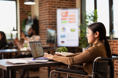 Businesswoman with a disability uses her laptop at desk, analyzing company growth charts and...