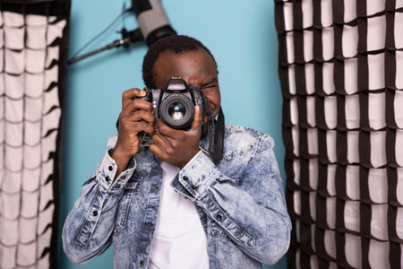 Black male photographer, deeply focused, using his DSLR camera in a studio setting. African american man positioned near softboxes and professional lighting, preparing to capture high quality images.の写真素材