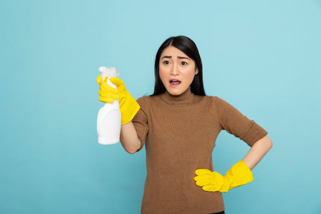 Surprised female cleaner in rubber gloves poses with spray bottle, pointing in disbelief. Asian housekeeper with eyes and mouth wide open, discovering messy house during morning housekeeping routine.の写真素材