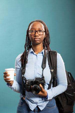 Female photographer standing with camera and disposable cup in her hands. African American female model poses confidently, wearing blue shirt, with DSLR around neck and holding coffee.の写真素材