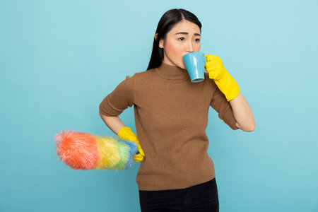 Asian female housekeeper holds a duster and enjoys coffee during spring cleaning. Pretty asian cleaner standing against blue background, taking relaxing break with mug to her mouth.の写真素材