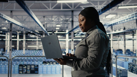 IT expert monitors server farm activity using laptop while walking in facility. African american woman in data center reviews rigs tests results to ensure systems are working correctly, camera Aの写真素材