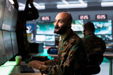 Army dispatcher looking at surveillance transmission on displays, relaying data to troops using headset microphone. Middle Eastern military operative transmitting reconnaissance infoの写真素材