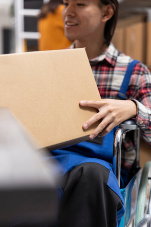 Male worker in wheelchair prepares packages for custom orders and local shipment. Home business logistics center supports chronic impairment with accessible shelving units and packaging station.の写真素材