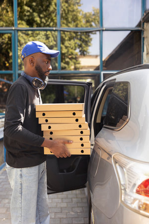 African american delivery man bringing a huge pizza order with many boxes, delivering fast food in the city by car. Male courier ensuring a quick service, drop off restaurant takeaway.の写真素材
