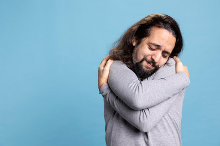 Young man practicing self love and self appreciation by hugging himself, being satisfied and fulfilled in studio. Powerful relaxed person shows self esteem and self care on camera.の写真素材