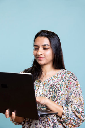 Young indian model holding a sleek laptop and doing freelancing tasks, typing on wireless computer...