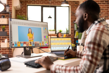 Focused male photo editor retouches images of female model on dual monitors in modern brick wall home. Creative tools, warm light and casual workspace sets productive, collaborative environment.の写真素材