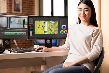 Asian female video editor posing confidently at home studio with color grading tools on dual...