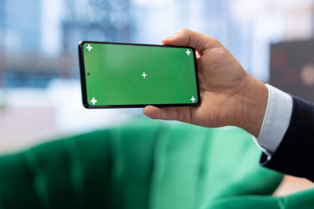 Determined male administrator uses phone with copy space display, tracking profit margins and making data driven decisions. Young entrepreneur working on a success story, multitasking.の写真素材