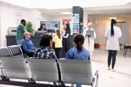 Hospital staff assist patients at front desk in organized emergency waiting area. Nurse in blue...