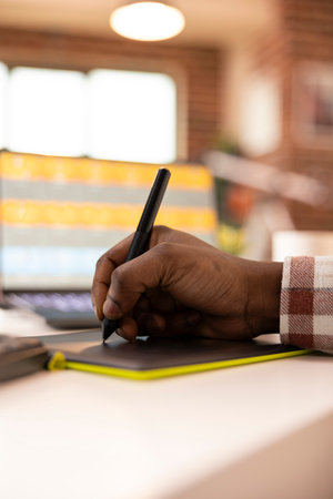 Close up of african american hand using stylus and graphic tablet to retouch images on laptop. Black...