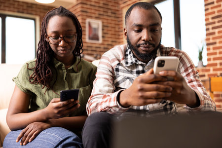 African american husband and wife sitting on couch, each focused on their mobile phones. Young black couple browsing social media and texting, enjoying digital lifestyle in brick wall apartment.の写真素材