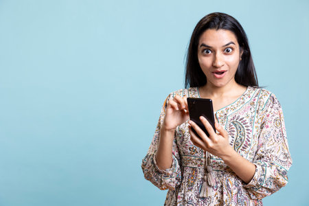 Surprised indian person using her mobile phone for social media apps and websites, browsing pages against blue background. Young woman being cheerful and amazed in the studio, smartphone.の写真素材