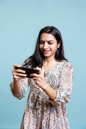 Ecstatic woman playing mobile video games on her smartphone, playing to defeat foes and clenching her hands. Indian female gamer enjoying leisure activity during her free time in the studio.の写真素材