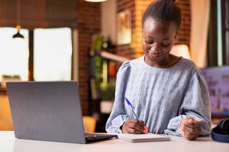 Focused female freelancer working indoors, reviewing sales performance on laptop and taking notes. Remote worker writing ideas in notepad while managing reporting and organizational tasks.の写真素材