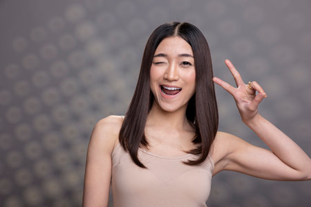 Asian studio model showing a peace sign and winking at the camera, feeling cheerful and confident in her skin. Flawless woman being feminine and adorable, body positivity and empowerment.の写真素材