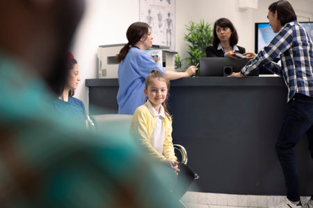 Portrait of caucasian girl waiting patiently in clinic lobby as asian man pays at reception desk. In...