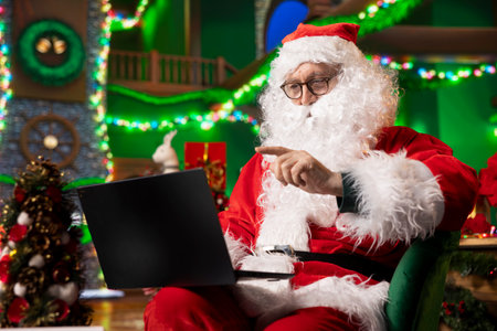Santa using laptop in festive decorated North Pole workshop to check heartfelt messages from kids...