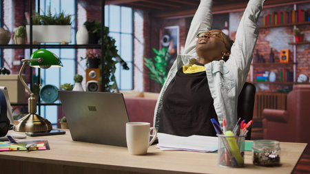Woman in home office stretches back, feeling fatigued while working on laptop. Remote employee in...