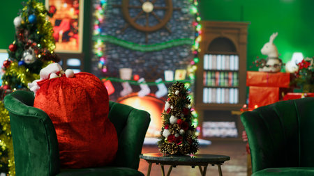 Small Christmas tree full of colorful garlands on table next to red Santa bag full of gifts. Cozy wintertime atmosphere in Santa Claus workshop adorn with holiday props, handheld camera shotの写真素材