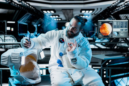 Astronaut takes selfie inside space station with Mars mission screen glowing in background. Starship crew member doing pictures using mobile phone before landing on red planet, making hand gesturesの写真素材