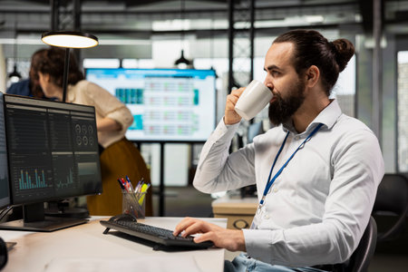 Employee sipping coffee while he reviews analytics and KPI metrics on monitor, driving business...