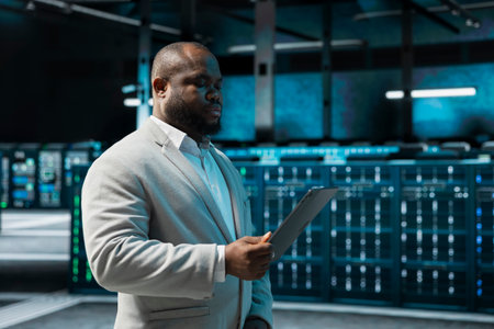 Programmer in data center updating systems using tablet, debugging app performance issues. Server hub IT expert using device, looking for malfunctions, increasing maximum efficiency and uptimeの写真素材