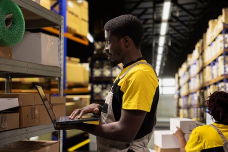 Black male staff verifying inventory pallets in e-commerce logistics center. Focusing on accurate order processing and packaging for quick shipping in large scale warehouse setting.の写真素材