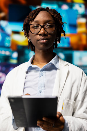 Doctor in lab coat monitoring medical research data and 3D anatomy models dashboards with tablet app. Clinician holding touchscreen device standing near multimedia wall showing visual medical reportsの写真素材