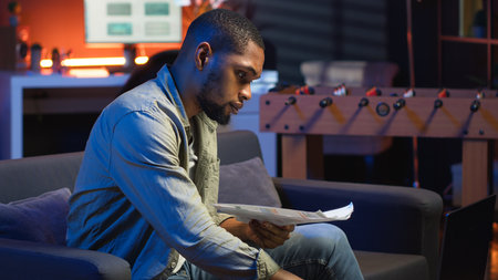 Black freelancer sitting on sofa and reading some papers to do research, multitasking with archives in the living room. Adult diving into professional or personal topics of improvement. Camera B.の写真素材