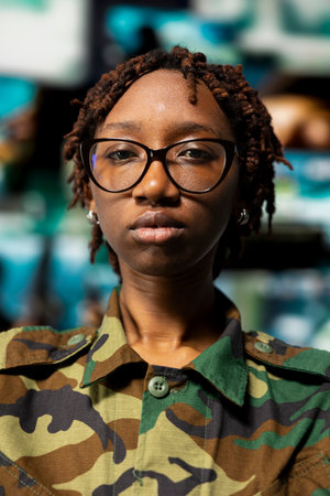 Closeup portrait of soldier in front of command center screens used for surveillance and threat monitoring. African american officer oversees mission control with displays showing intelligence feedsの写真素材