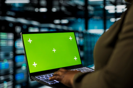 Programmer uses green screen mockup laptop to do maintenance in data center with setup helping companies stay connected. Worker walks with chroma key notebook between server hub rigs blinking lightsの写真素材