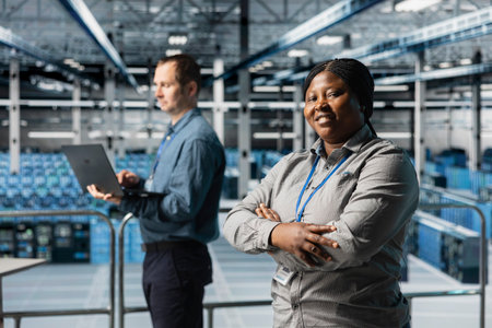 Portrait of smiling server room engineer next to coworker troubleshooting errors using software on laptop. Cheerful african american woman in data center and colleague fixing bugs using notebookの写真素材