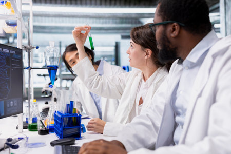Multiethnic team of lab technicians inspect chemical solution clarity in vials for purity verification. Research facility workers brainstorming, preparing reagent test tubes for assay proceduresの写真素材