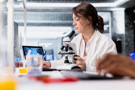 Scientist using microscope to evaluate cellular structures in lab workspace, writing down logs. Woman observes specimens through magnification tool at laboratory bench workstation, taking notesの写真素材