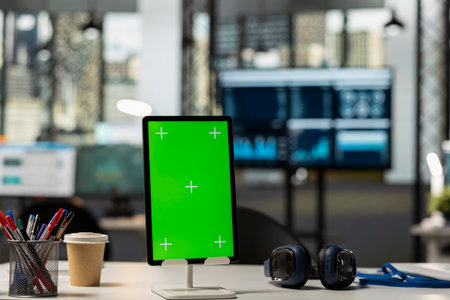 Empty modern office desk with tablet displaying green screen template. Digital device on table in vacant workplace with isolated copy space for financial content or company branding.の写真素材