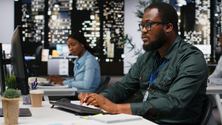 African american office workers solving tasks after hours at workplace, typing information on desktop pc to create a new report for performance data. Finishing project late at night.の写真素材