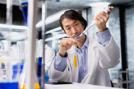 Senior scientist uses pipette to transfer liquid into test tube containing yellow solution at laboratory bench. Asian man does pipetting procedure with chemical liquid solution in test tubeの写真素材