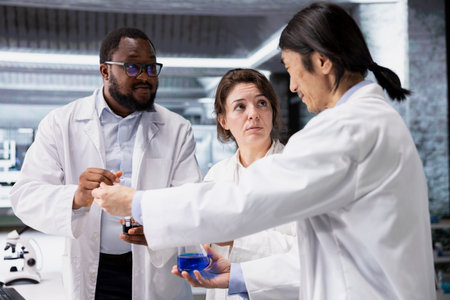 Team of lab chemists inspecting clarity of chemical solution in glass flasks for purity verification. Multiethnic research facility colleagues prepare reagent containers for assay proceduresの写真素材