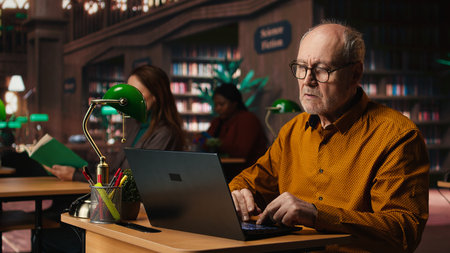 Elderly male adult reviewing academic text on a laptop at the university, sitting in classic study area at library and browsing online documents. Education through formal learning.の写真素材