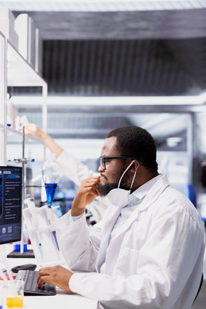 Lab scientist wearing protective face mask evaluates genetic markers associated with disease risk using PC. Expert wearing medical mask studies DNA strand mapping with genomics analysis programの写真素材