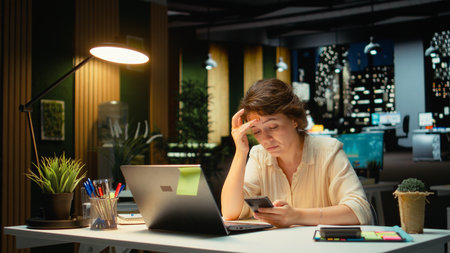 Weary businesswoman stays connected on social media while she does overtime at night, checking her mobile phone after receiving a notification. Female worker checks text messages. Camera A.の写真素材