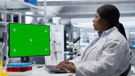 Green screen desktop next to black woman researcher performs bio science in modern laboratory, using pc and conducting an experiment for biotechnology analysis. Clinical science.の写真素材