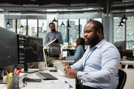 Trader at work tracking stock exchange metrics and placing orders using trading software on PC. African american man in brokerage company office trading securities on behalf of clientsの写真素材