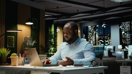 Black businessman sighing and checking messages on mobile phone, feeling drained and frowning after being distracted by a notification in modern office. Workplace challenges at night.の写真素材
