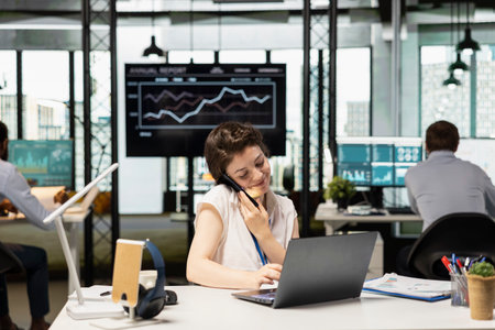 Smiling caucasian office worker multitasks, talking on mobile phone and using laptop at desk. Female...