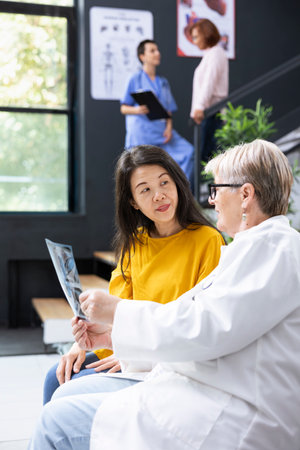 During a friendly consultation, elderly doctor explains radiology scan results to asian woman. Hospital specialist ensuring healthcare access and professionalism in treating sick people.の写真素材