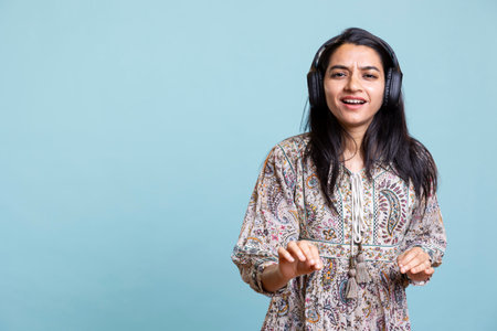 Happy woman playing electronic music and dancing around on camera, pretending to use turntables. Indian person feels joyful wearing headphones and making songs over studio background.の写真素材