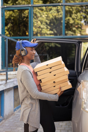 Black takeaway employee parking the car to bring pizza boxes to people at the doorstep, delivering a massive fast food order. Female courier ensuring an express five star service.の写真素材